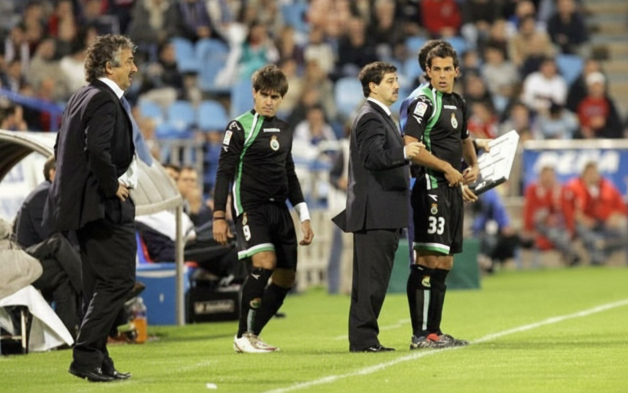 Dos jugadores del Racing de Santander, con los dorsales 9 y 33, están en la banda listos para entrar al campo. Están acompañados por dos entrenadores con traje. Al fondo se ven gradas con público.